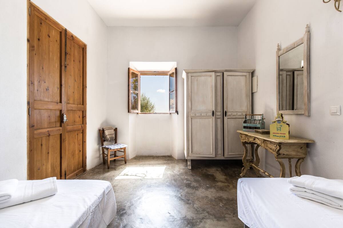 A room with two beds, a wooden wardrobe, a small table with a mirror, and a window with a view of trees.