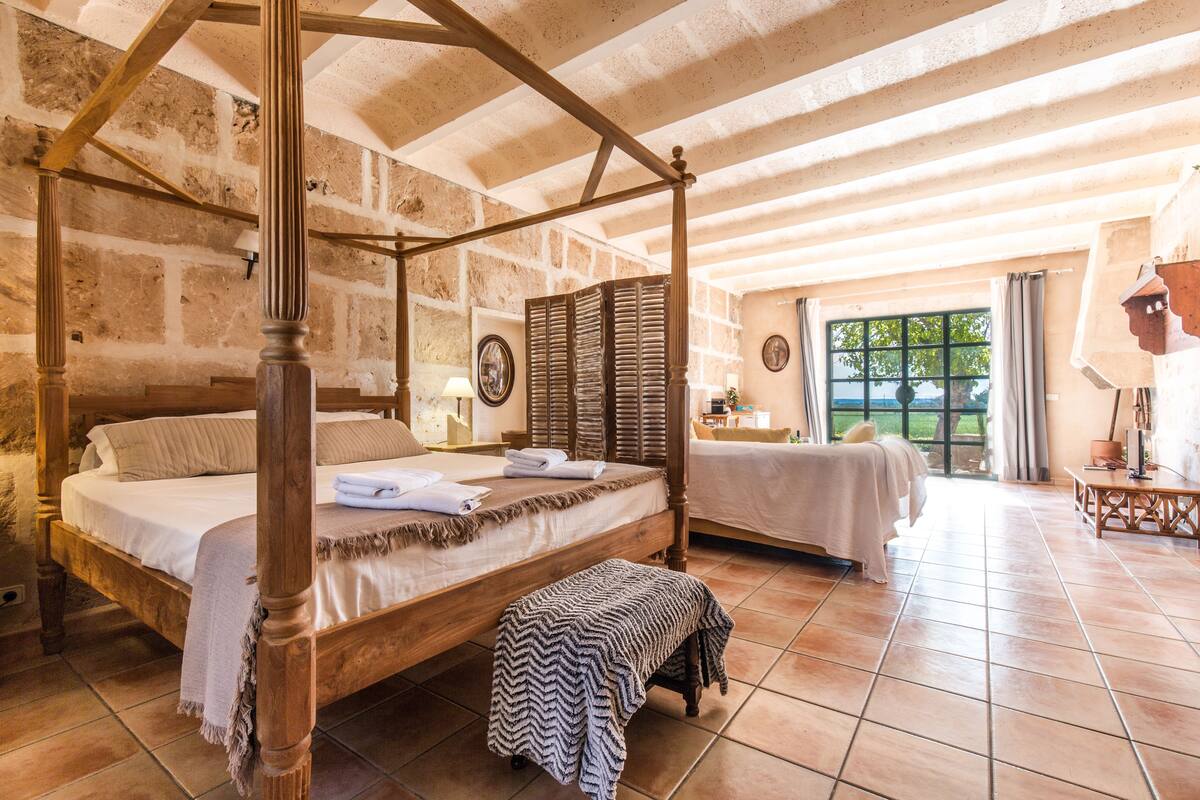 A four-poster bed with a bench, a bedside table, and a window with curtains.