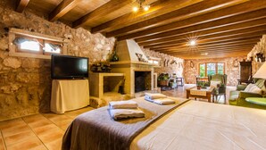 Una habitación rústica con pared de piedra, vigas de madera, chimenea, televisor de pantalla plana, cama con toallas, mesa de comedor y un sillón verde.