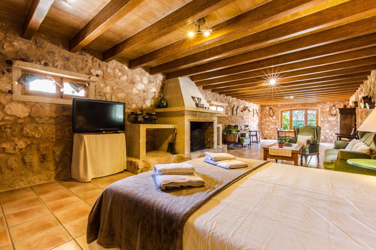 A rustic room with a stone wall, wooden beams, a fireplace, a flat-screen TV, a bed with towels, a dining table, and a green armchair.
