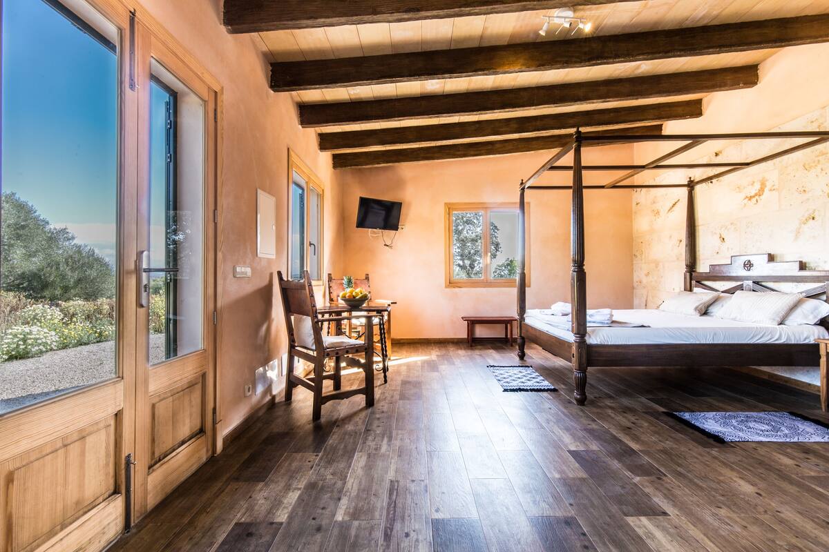A bedroom with a wooden floor, a four-poster bed, a television on the wall, and a window with a view of trees.