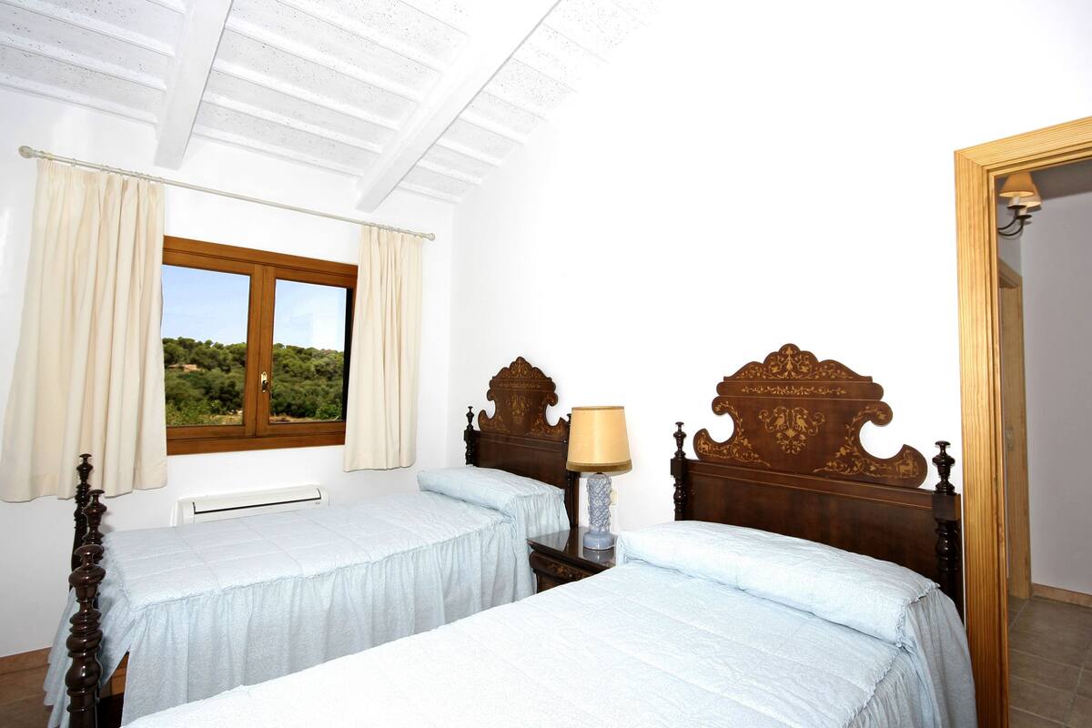 A bedroom with two beds, a window with curtains, a nightstand with a lamp, and a wooden headboard.
