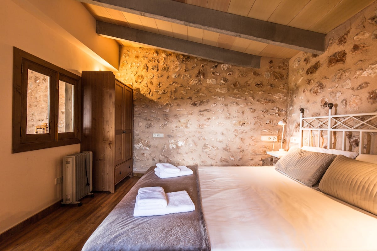 A bedroom with a stone wall, wooden beams, a bed with pillows, a radiator, and a window.