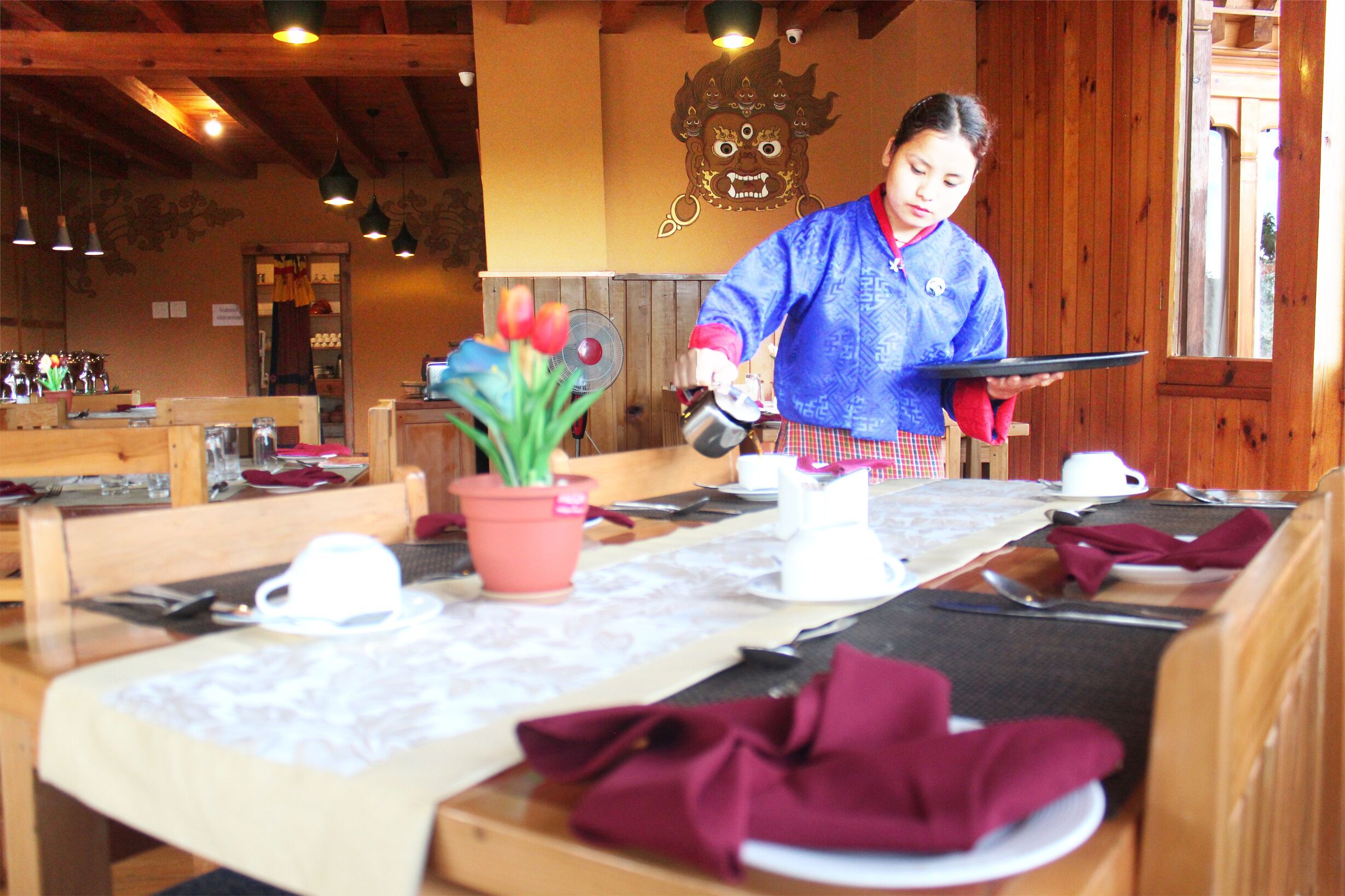 A restaurant interior with wooden tables set for dining, a potted plant on the table, and a person serving food.