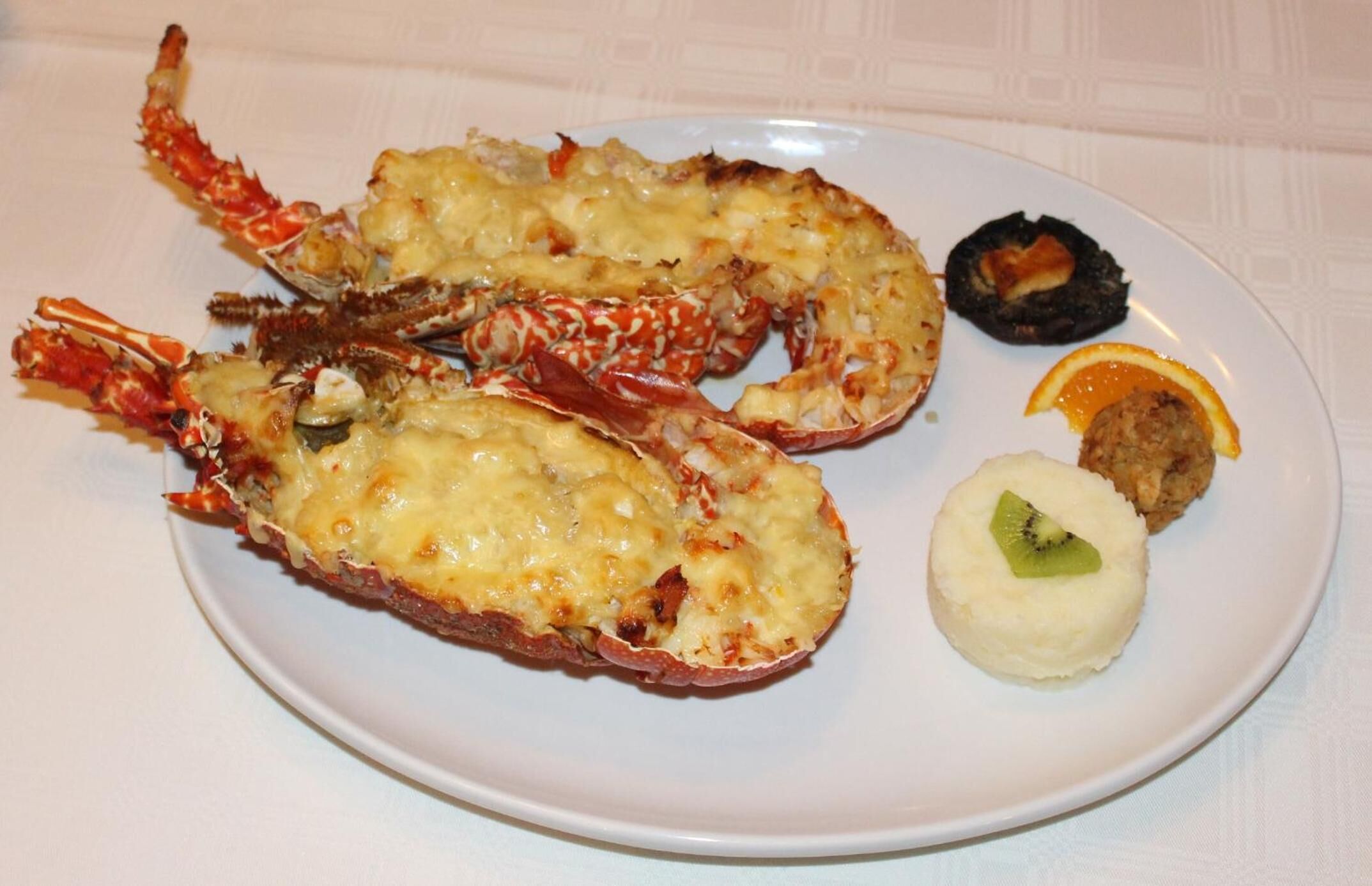 Une assiette avec deux homards cuits, une tranche d’orange et une portion de purée de pommes de terre garnie d’une tranche de kiwi.