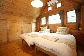 Dormitorio de cabaña de madera con dos camas, un ventilador de techo y ventanas grandes con cortinas.