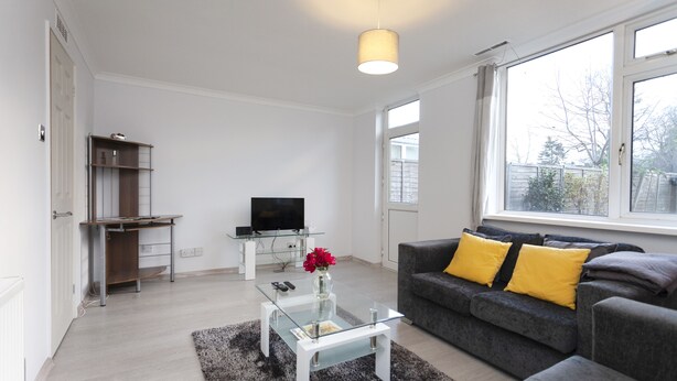 Ein modernes Wohnzimmer mit Glastisch, grauem Sofa mit gelben Kissen, Flachbildfernseher auf einem Standfuß und einem großen Fenster mit Blick ins Freie.