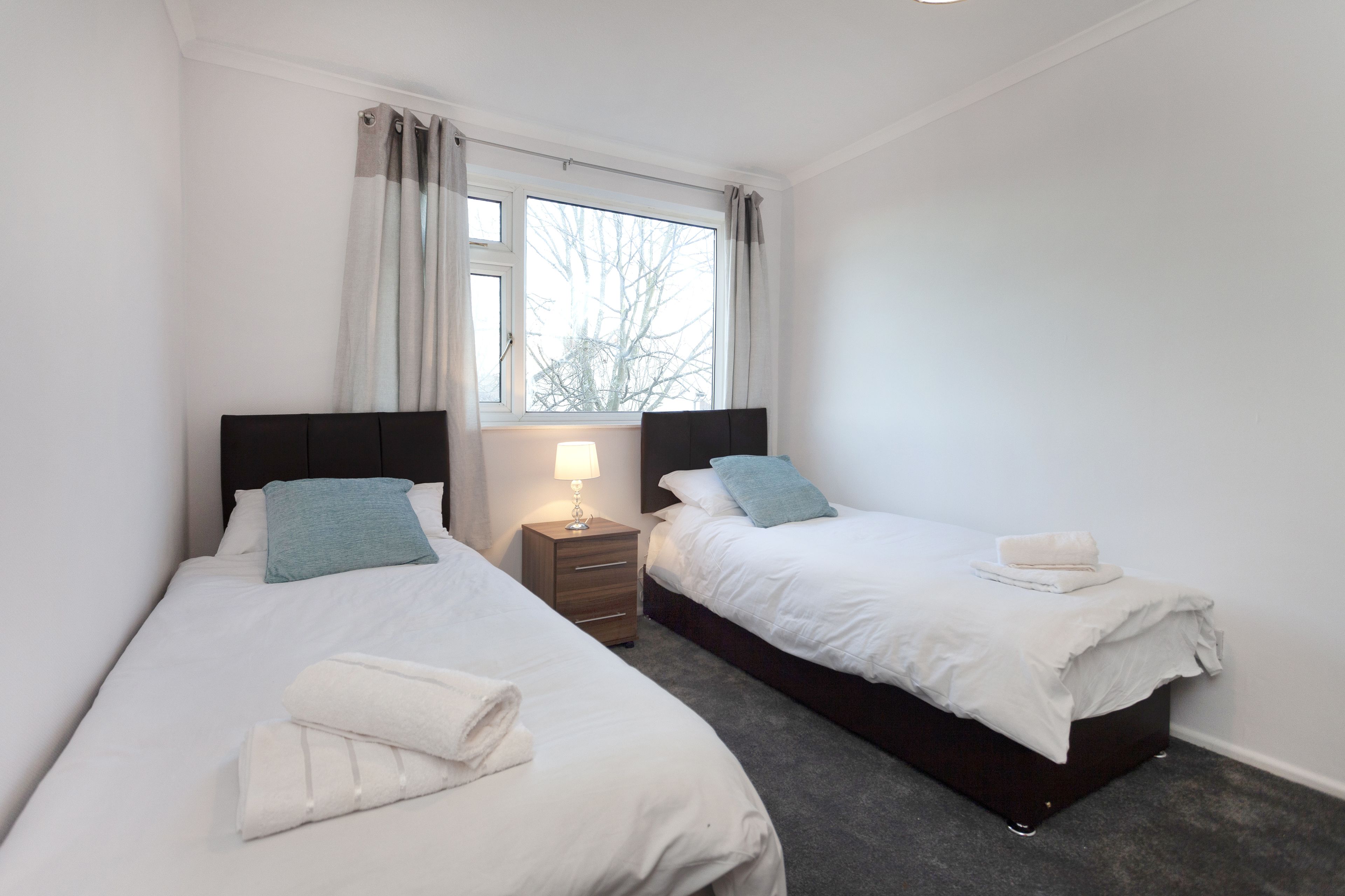 A hotel room with two single beds, a nightstand with a lamp, and a window with curtains.
