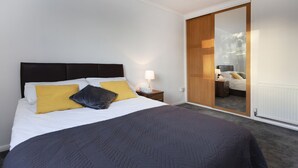 Ein ordentlich bezogenes Bett mit dunklem Kopfteil, ein Nachttisch mit Lampe und ein großer Spiegel, der ein Schlafzimmer mit Bett und Kommode reflektiert.