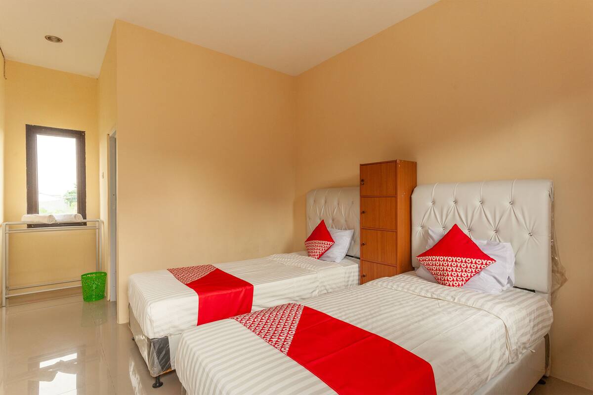 A hotel room with two beds, a wooden wardrobe, a window, and a green trash bin.