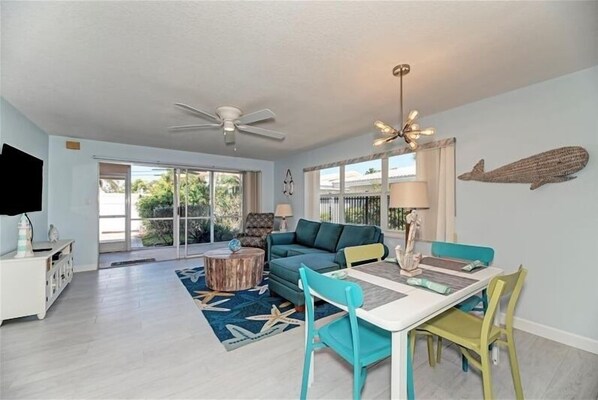 A spacious living area with a sofa, dining table, and chairs, a ceiling fan, and a large window with a view of greenery.