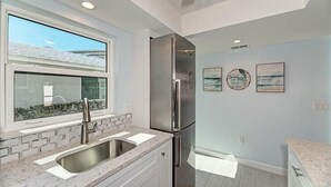 A kitchen with a stainless steel refrigerator, a sink with a marble countertop, and a window overlooking a snowy landscape. - Crystal Sands Villa 12 - Private Beach (Sarasota)