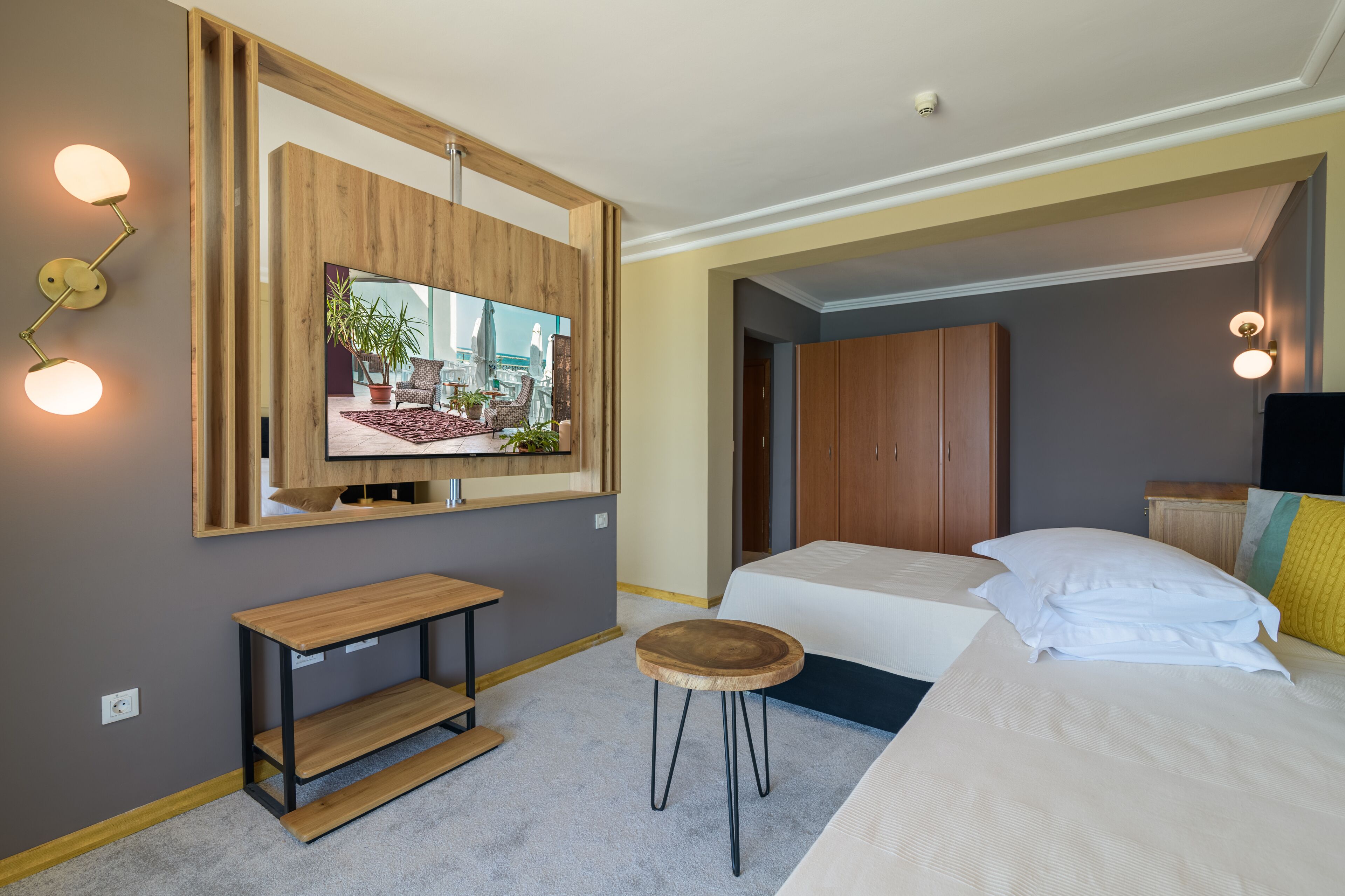 A hotel room with a large mirror, a television, a wooden bench, a round stool, two beds, and a wardrobe.