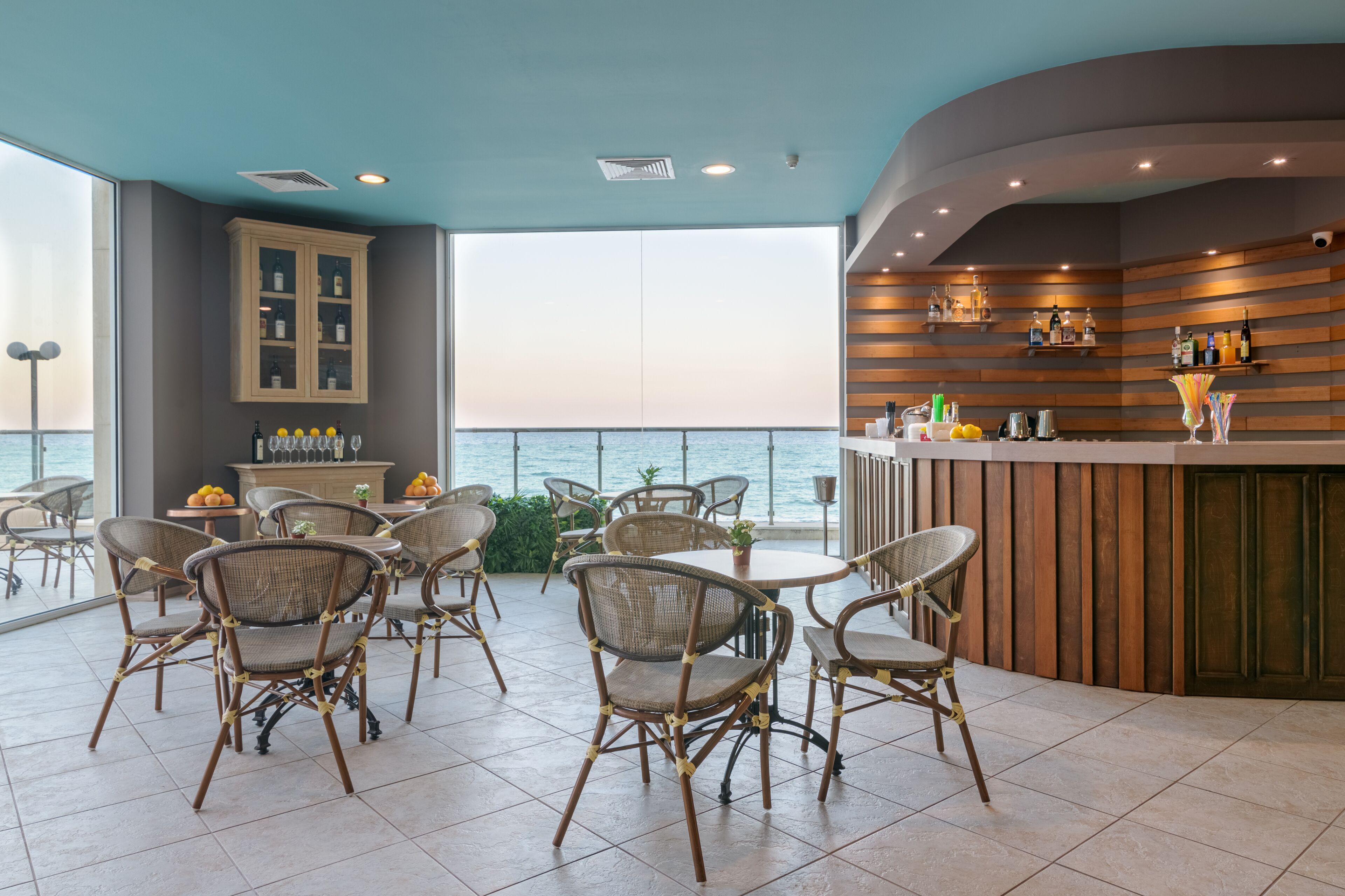 A spacious outdoor seating area with wicker chairs and tables, a bar with wooden shelves, and a view of the sea.
