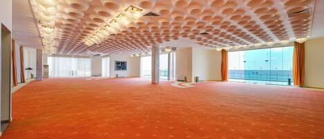 A spacious room with a patterned carpet, a unique ceiling design, and large windows offering a view.