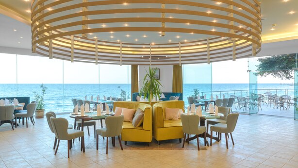 A modern dining area with a yellow sofa and chairs, a central plant, and a view of the sea. - Marina Sunny Day Resort (Varna)