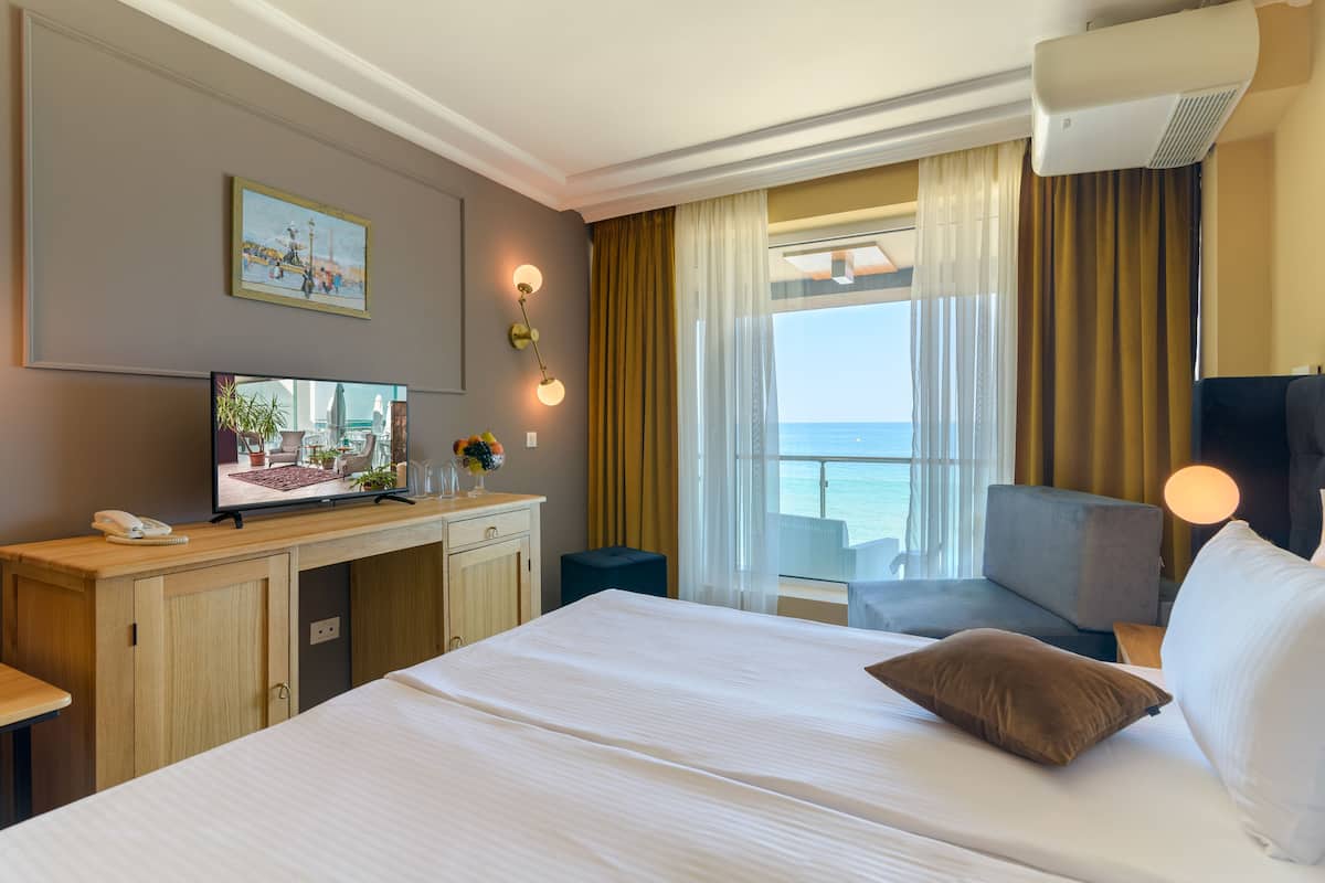 A hotel room with a large bed, a desk with a computer, a television, and a view of the sea.