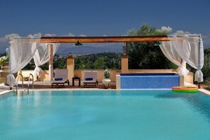 A pool area with a wooden pergola, white drapes, and a view of a mountainous landscape.