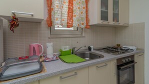 A kitchen with a sink, stove, and cabinets. There is a window with a curtain, a kettle, and a dish rack. - Flower House (Kastela)