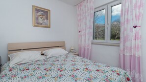 A bedroom with a bed, a window with curtains, a nightstand, and a wall-mounted picture. - Flower House (Kastela)