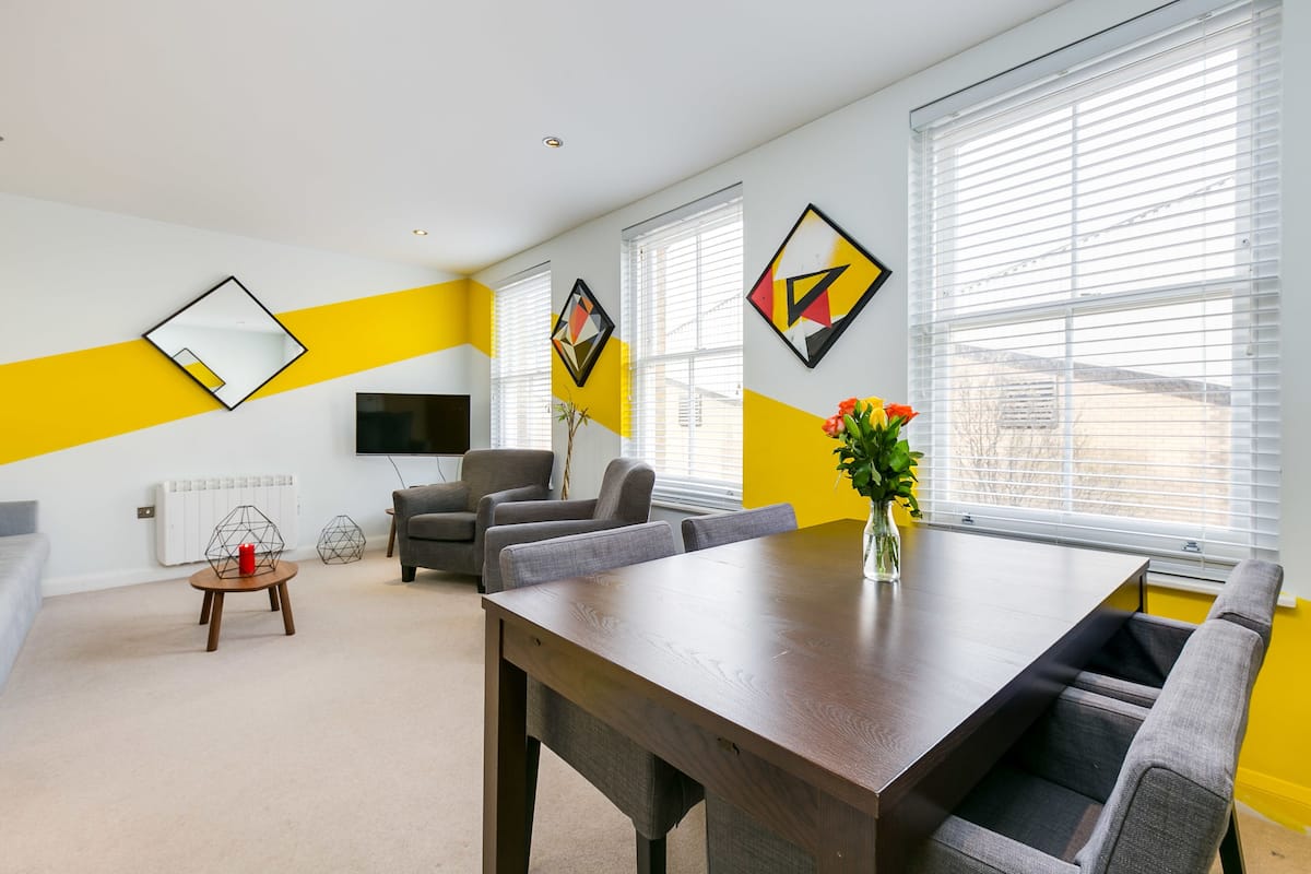 A modern living room with a wooden dining table, grey sofas, and a TV mounted on the wall.