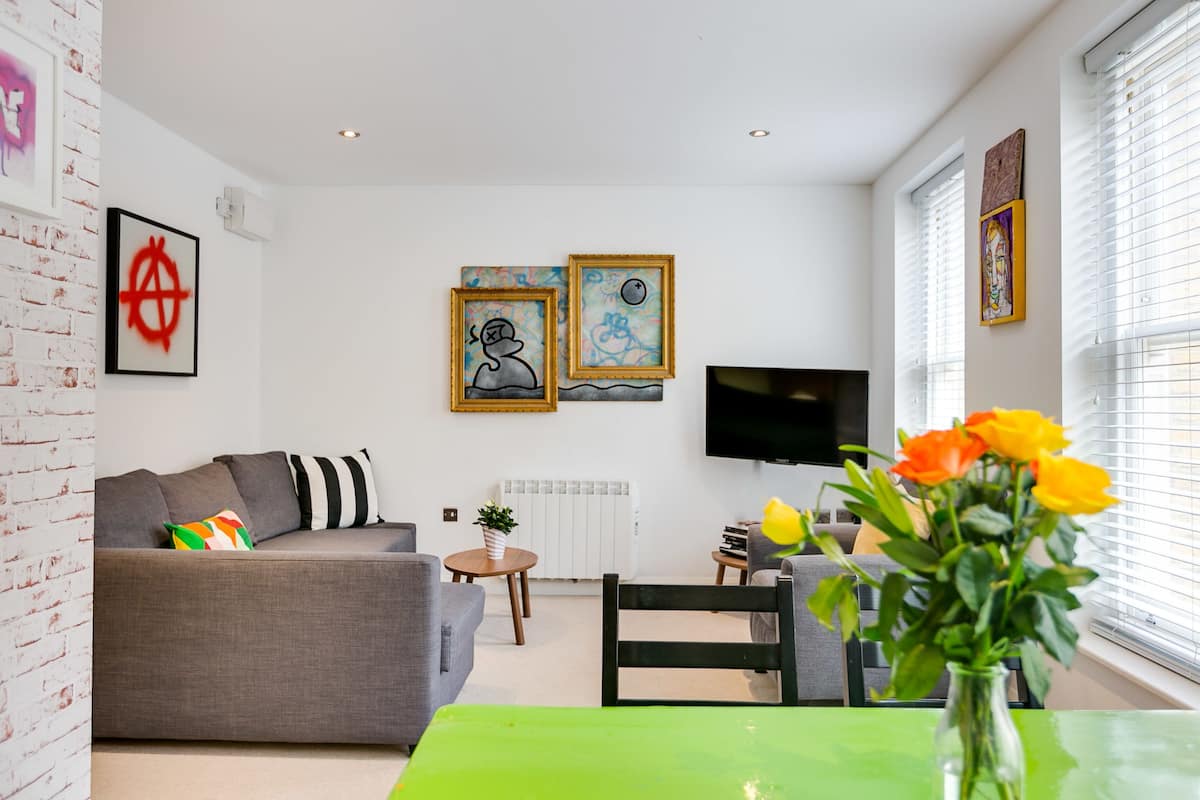 A modern living room with a grey sofa, a green table, and a TV. There are two framed artworks on the wall and a window with blinds.