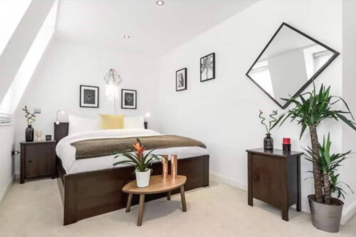 A bedroom with a bed, bedside tables, a small round table with a plant, a mirror, and framed pictures on the wall.