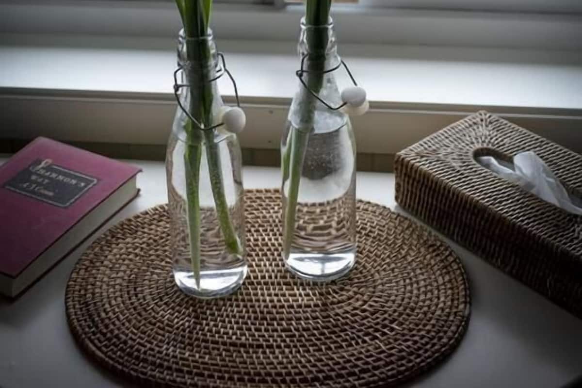 Two glass vases with purple flowers on a woven mat by a window.