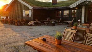 Un edificio rústico de madera con techo inclinado, una chimenea y un patio de grava con mesas y sillas de madera.