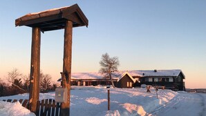 Una estructura de madera con un buzón, un paisaje cubierto de nieve y un edificio con techo inclinado al fondo.