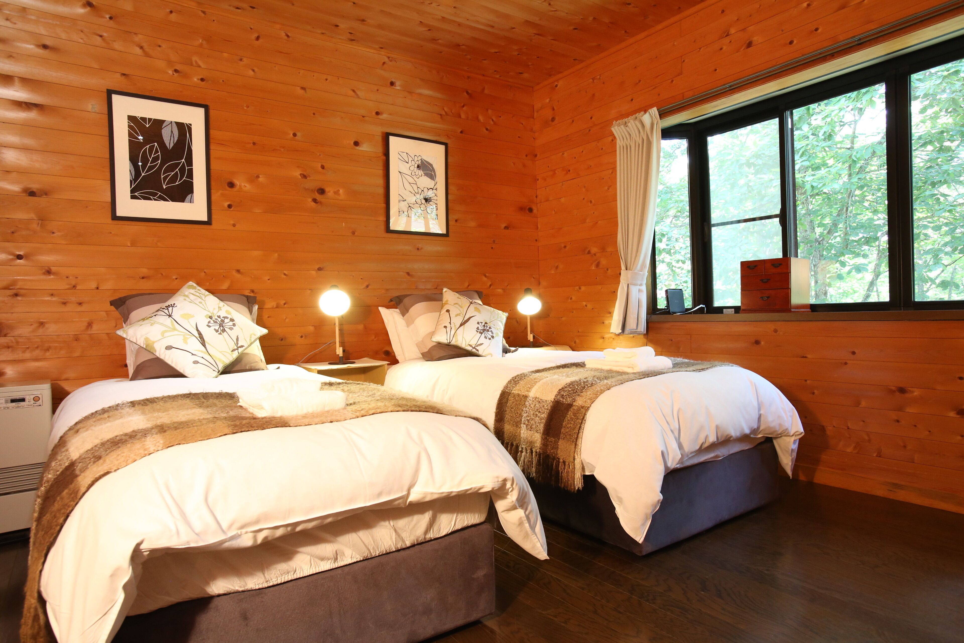 A wooden cabin bedroom with two beds, framed artwork on the walls, and a window with curtains.