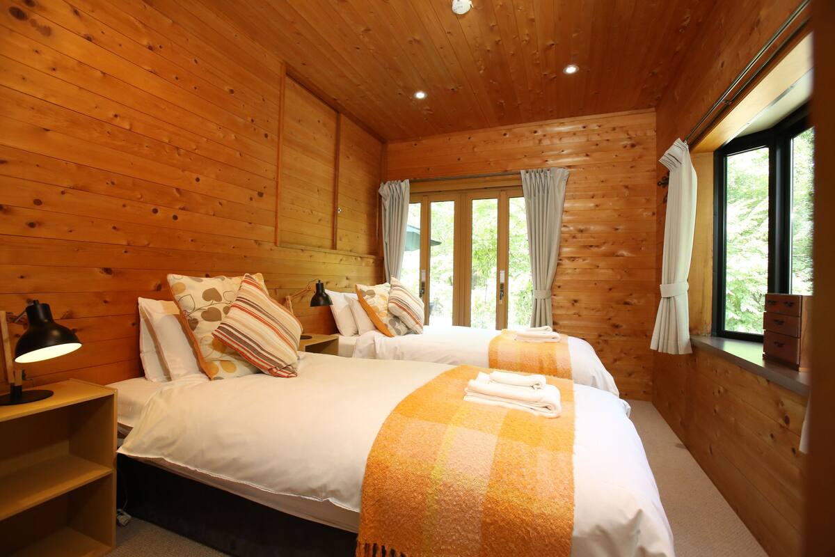 A wooden cabin room with two beds, a nightstand, a lamp, and a window with curtains.