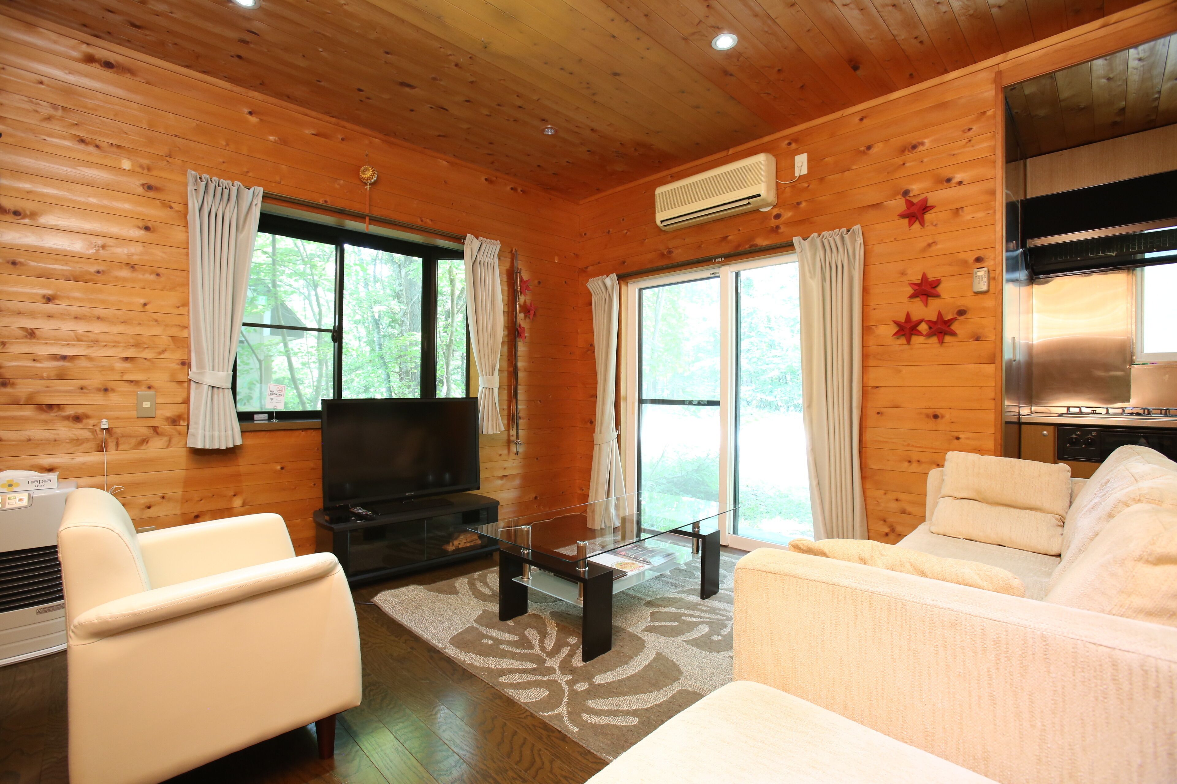 A wooden cabin interior with a sofa, a glass coffee table, a television, and a kitchen area.