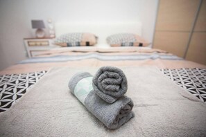 A neatly made bed with a rolled towel in the center, a patterned bedspread, and a bedside table with a lamp and a small decorative item.