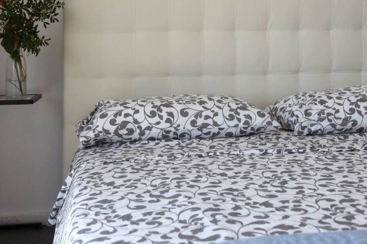 Une chambre à coucher avec une tête de lit capitonnée, une couvre-lit à motifs et un vase rempli de fleurs posé sur une table de chevet.