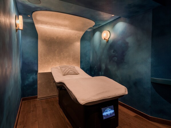 Een massageruimte met een massagetafel, een aan de muur bevestigde massaglamp en een scherm waarop een afspeellijst voor massages wordt weergegeven.
