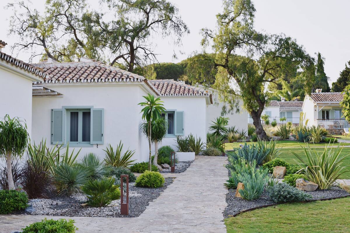 A white building with a tiled roof and green shutters, surrounded by a well-maintained garden with various plants and a stone pathway.