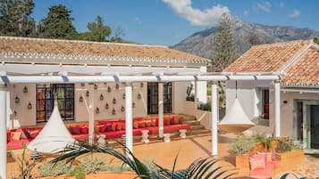 Un espacio de descanso al aire libre con cojines rojos y beige, rodeado de plantas en macetas y vistas a montañas.