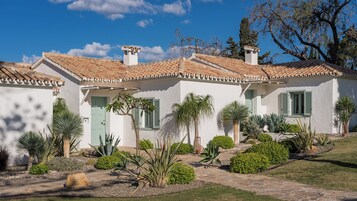 Un edificio blanco con techo de tejas, contraventanas verdes y un pequeño jardín con diversas plantas.