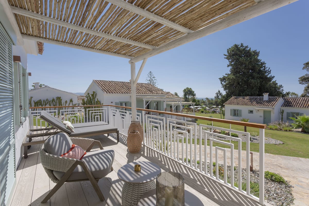 A covered outdoor area with lounge chairs and a small table, surrounded by a white fence and a view of houses and trees.