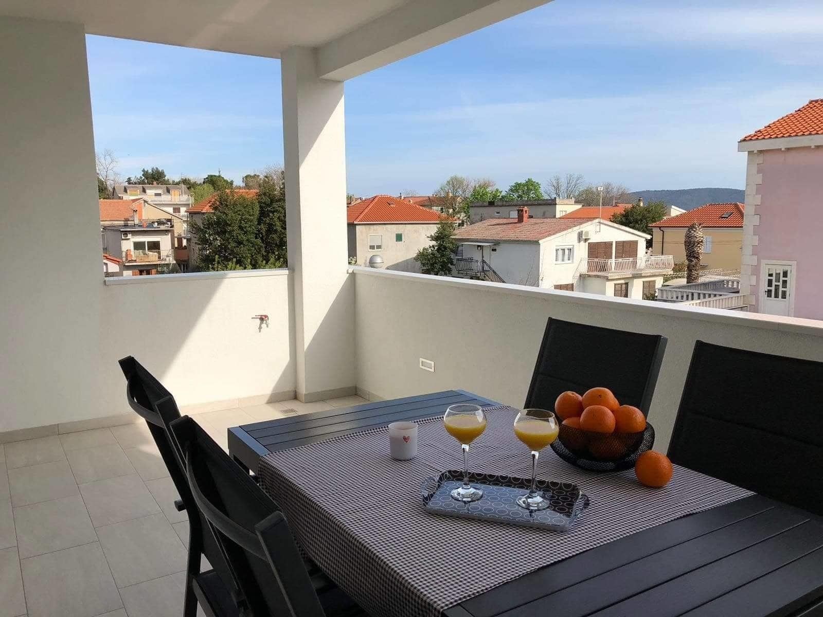 Ένα μπαλκόνι με ένα τραπέζι στρωμένο για δύο άτομα, στο οποίο υπάρχει ένα μπολ με πορτοκάλια και ποτήρια με χυμό πορτοκαλιού.
