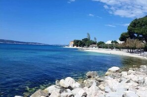 Blick auf die Küste mit klarem, blauem Wasser, felsigem Ufer und Gebäuden entlang des Strandes.