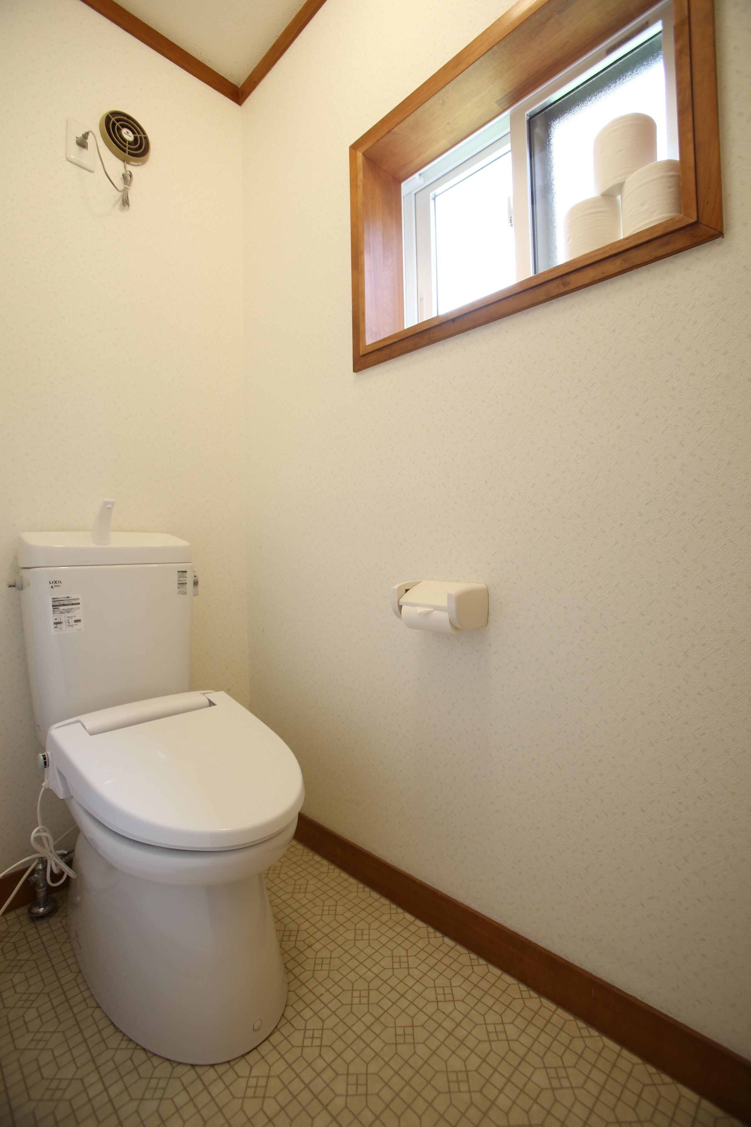 Un baño con inodoro, dispensador de papel higiénico en la pared, ventana con marco de madera y ventilador fijado a la pared.