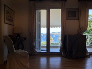 A room with a wooden floor, a chair, a table, and a view of a balcony with a railing and a body of water. - Luxury Apartment Sea View Garden Parking (Corfu)