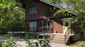 A two-story cabin with a porch, large windows, and a staircase leading to the entrance. - Alps Aunty Cottage (Hakuba)
