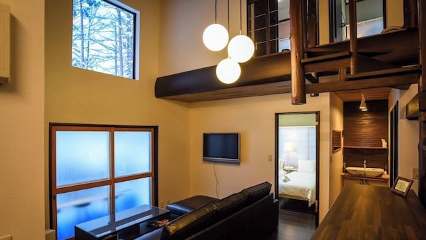 A modern living room with a sofa, a TV, a large window, and a staircase. - Alps Aunty Cottage (Hakuba)