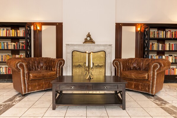 A room with two leather armchairs, a dark wooden coffee table, and a fireplace with a clock above it. Bookshelves line the walls.