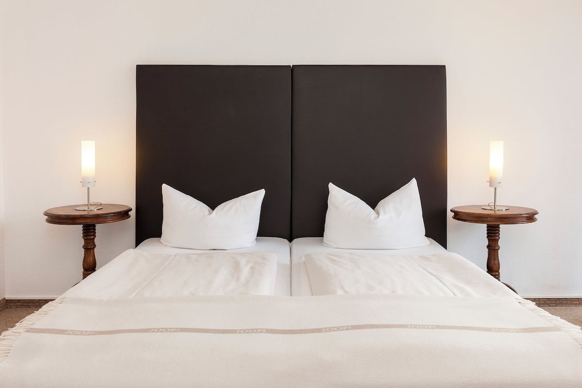 Een tweepersoonsbed met een zwart hoofdeinde, twee witte kussens en een witte sprei. Aan weerszijden van het bed staan twee nachtkastjes met lampen.