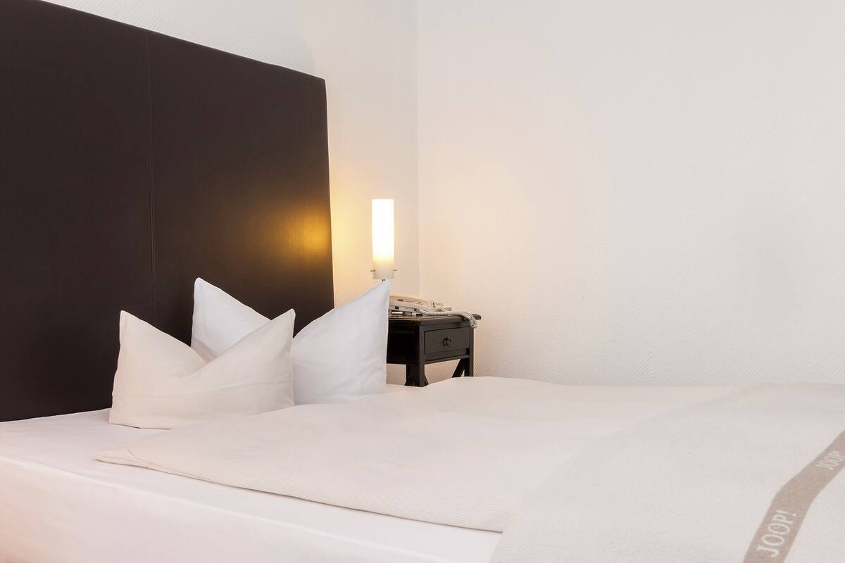 Een netjes opgemaakt bed met wit bedgoed en kussens, een nachtkastje met een lamp, en een donkere hoofdbord.