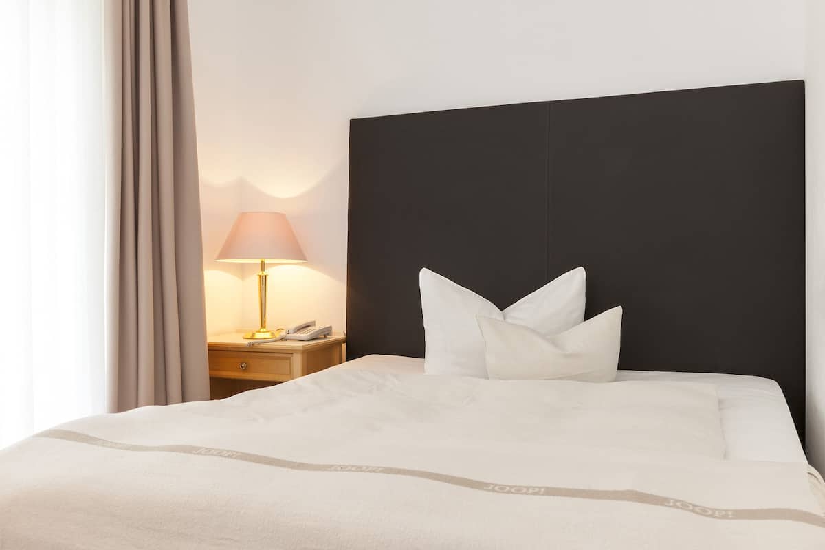 Een net opgemaakt bed met witte lakens en kussens, een bedlampje en een nachtkastje met een telefoon.